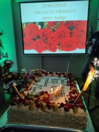 Na pierwszym planie duży tort ze świeczkami, w tle prezentacja na rzutniku "Jubileusz 100-lecia urodzin Pani Marii"